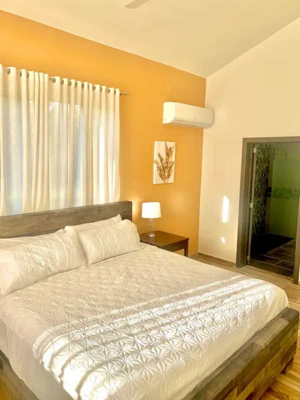 A neatly arranged bedroom featuring an orange wall, a large bed with white bedding, a wooden headboard, a lamp on a bedside table, and white curtains.