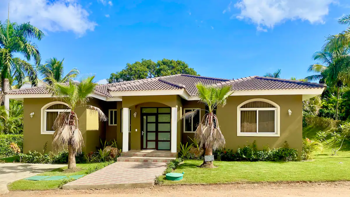 A single-story beige house with a brown tiled roof, surrounded by palm trees and a well-manicured lawn, under a clear blue sky.