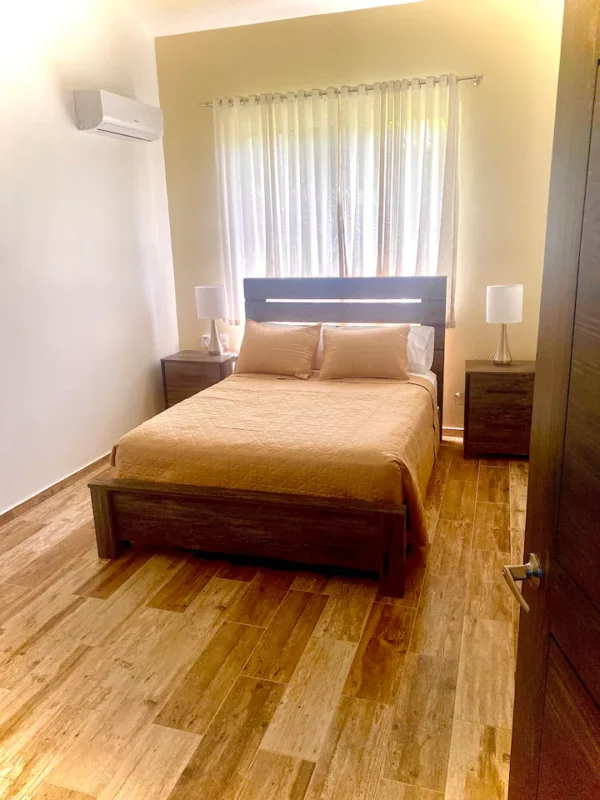 A modern bedroom with a large double bed, wooden floor, white curtains, and two bedside lamps.