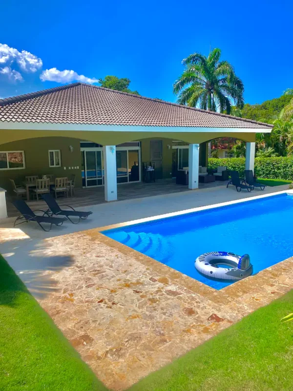 Luxury house with a large swimming pool, lounge chairs, inflatable ring, and lush greenery under a clear blue sky.