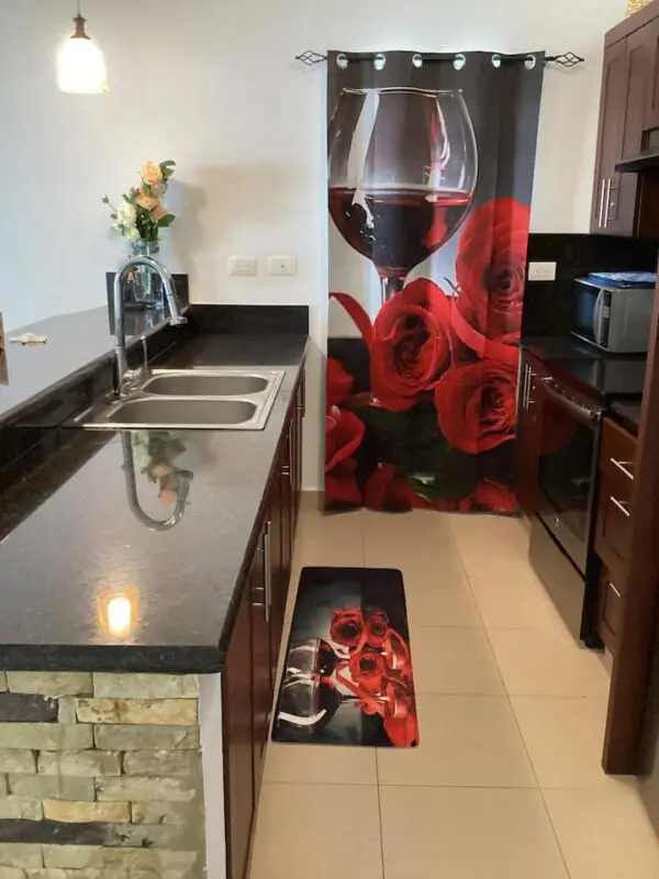 A modern kitchen with Ocean Front Lavish *Jr Penthouse* Exquisite Views, dark wood cabinets, a shiny black countertop, stainless steel appliances, and decorative curtains and rug featuring red roses and a wine glass design.