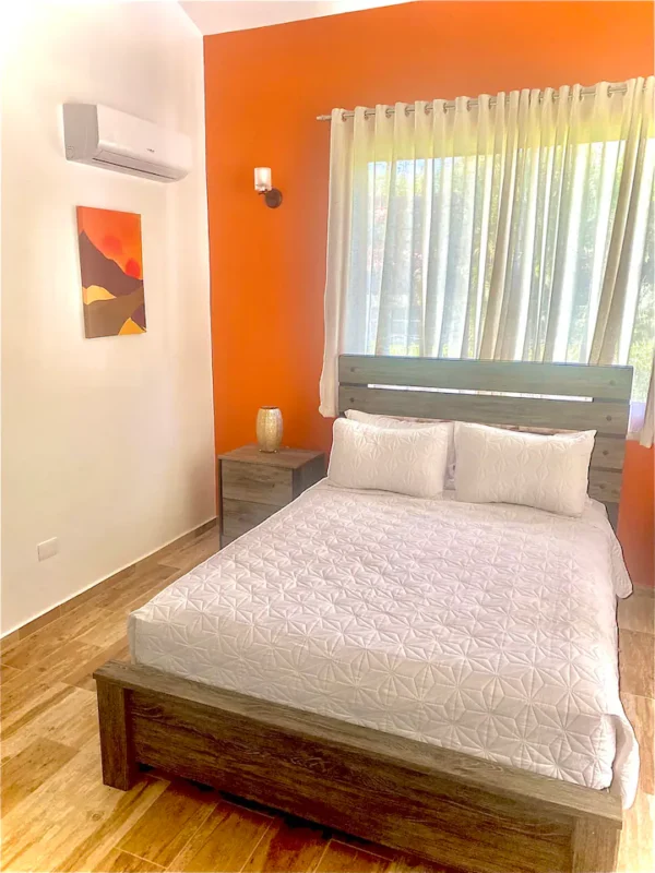 A modern bedroom with a double bed, orange walls, wooden headboard and frame, white bedding, and a wall-mounted air conditioner.