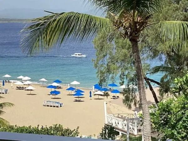 View from Ocean Front Lavish *Jr Penthouse* Exquisite Views overlooking a tropical beach with blue umbrellas, white lounge chairs, a palm tree, and a boat on the clear blue water.