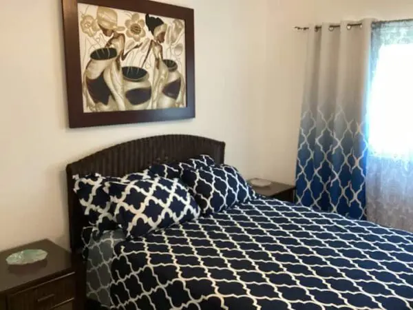 A tidy bedroom featuring a bed with Ocean Front Lavish *Jr Penthouse* Exquisite Views bedding, a framed abstract art piece above the bed, and a window with patterned curtains.