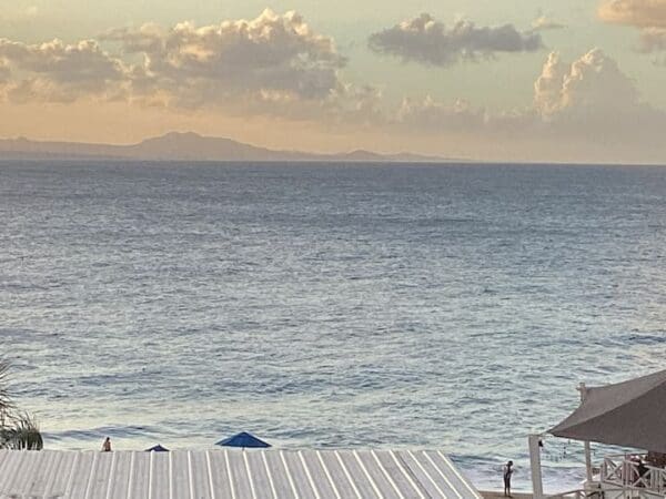 Ocean Front Lavish Jr Penthouse Exquisite Views with distant mountains, a white rooftop with two blue umbrellas, and people on a beach at sunset.