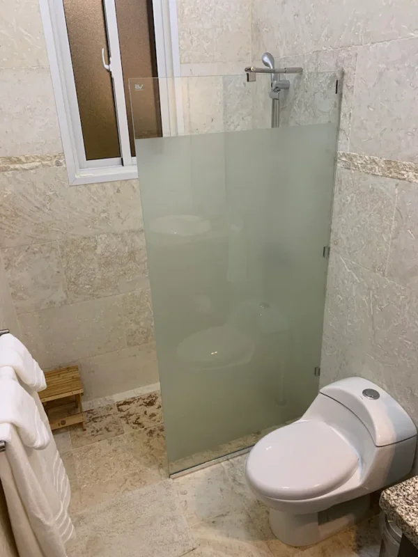 A modern bathroom with a frosted glass shower partition next to a white toilet, featuring beige stone walls and a small window.