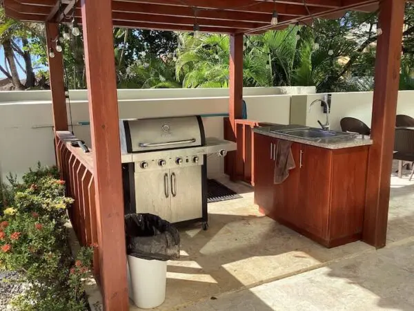 Outdoor kitchen with a Ocean Front Lavish *Jr Penthouse* Exquisite Views, sink, wooden cabinetry, and a trash can under a pergola, surrounded by lush greenery.