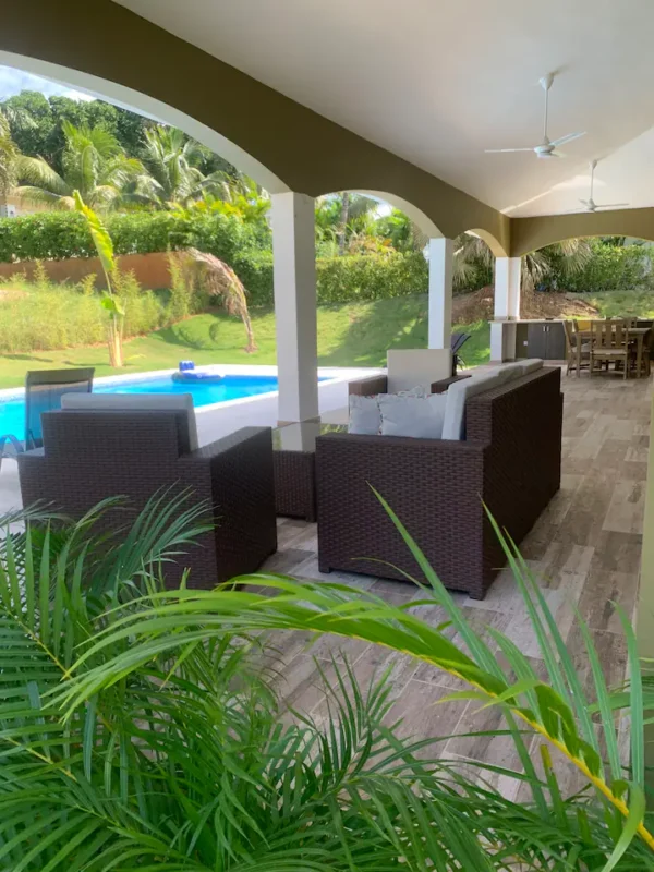 Covered patio with wicker furniture overlooking a swimming pool surrounded by lush greenery.