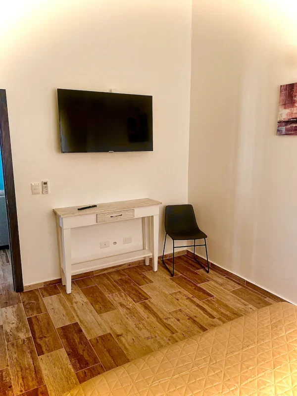 A modern room with a flat-screen TV on a white wall, a small white desk with drawers, and a black chair on a patterned wooden floor.