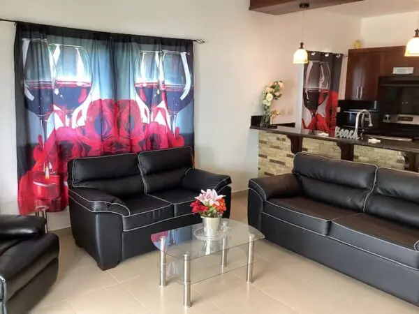 Modern living room with two black leather Ocean Front Lavish *Jr Penthouse* Exquisite Views, a glass center table, vibrant red floral curtains, and a small kitchen area in the background.