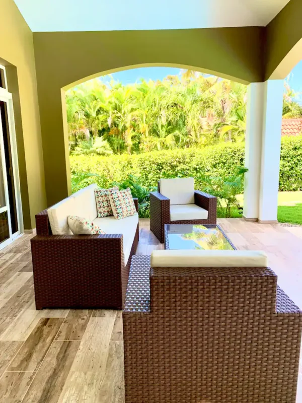 View of a covered patio with wicker furniture, including a sofa and chairs, overlooking a lush garden.