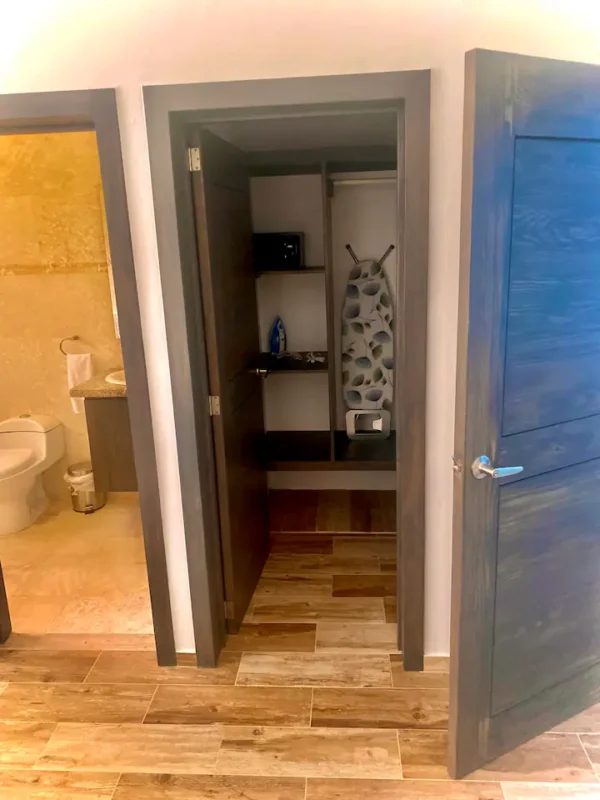 A bathroom with an open door leading to a closet housing a safe and ironing supplies, set on a wooden floor with beige walls.