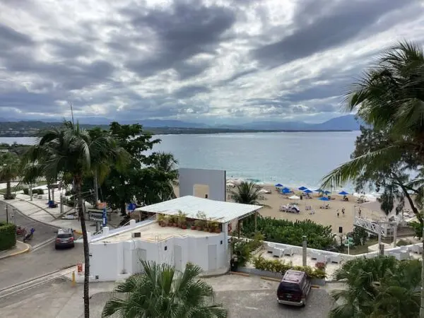 View of a Ocean Front Lavish *Jr Penthouse* Exquisite Views with scattered palm trees, a beach hut, and people on loungers under umbrellas, overlooking a calm bay with mountains in the distance.