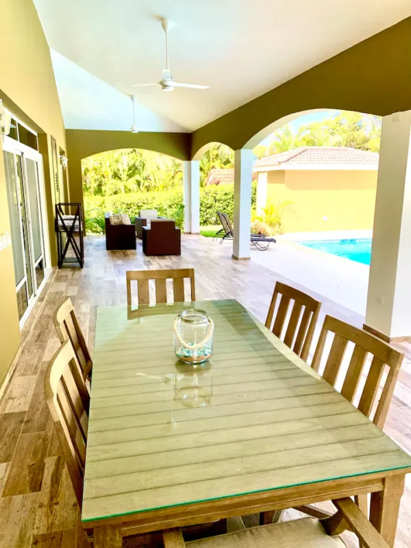 Spacious covered patio with a wooden dining table and chairs, overlooking a pool, in a sunny, tropical setting.