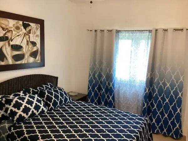 A neatly arranged Ocean Front Lavish Jr Penthouse with a blue and white patterned bedspread, wicker headboard, wall art depicting vases, and a window with blue patterned curtains.