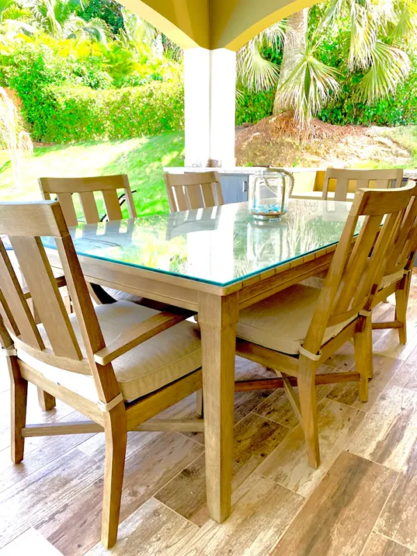 Outdoor dining set with a glass table and wooden chairs under a patio umbrella, surrounded by lush greenery.