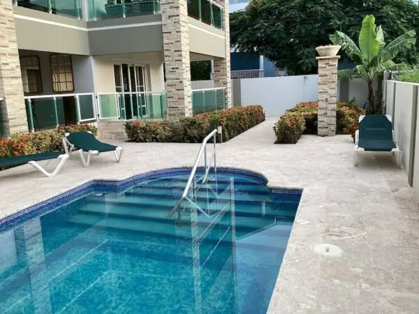 A backyard with a small blue Ocean Front Lavish *Jr Penthouse* Exquisite Views swimming pool, surrounded by a patio and plants, near a residential building. Two lounge chairs are placed beside the pool.