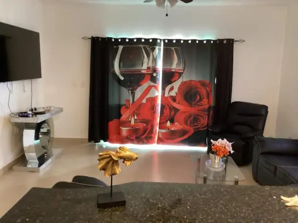 A living room featuring a large Ocean Front Lavish *Jr Penthouse* Exquisite Views curtain with a printed image of wine glasses and red roses, flanked by a black sofa, gaming console, and a glass coffee table with decorative items.