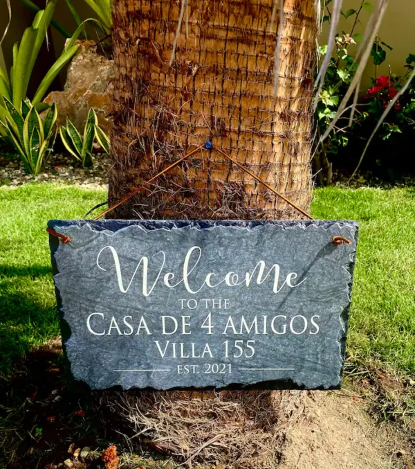 A slate welcome sign reading "Welcome to the Casa de 4 Amigos Villa 155 Est. 2021" hangs on a palm tree in a garden.