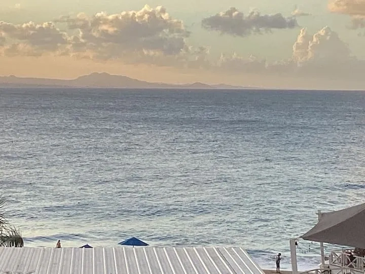 Ocean view with distant mountains, beachfront condos, and blue beach umbrellas, with people by the shore at sunset.