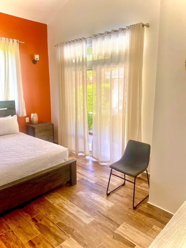 Brightly lit bedroom with orange walls, wooden floor, a large bed, sheer curtains, and a simple black chair.