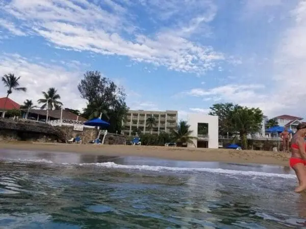 A beach scene with gentle waves in the foreground, a beachgoer on the right, and Ocean Front Lavish *Jr Penthouse* Exquisite Views with palm trees and blue umbrellas in the background under a cloudy sky.