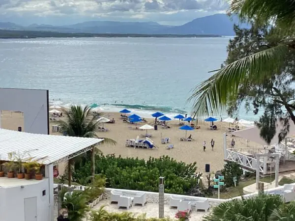 View of a beach with Ocean Front Lavish *Jr Penthouse* Exquisite Views umbrellas and loungers, adjacent to buildings, with mountains in the distance under a cloudy sky.