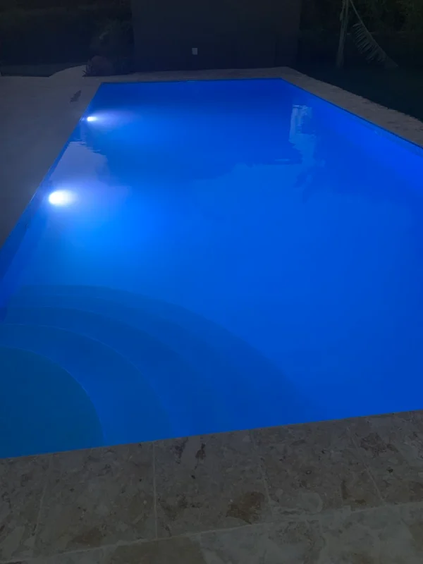 A swimming pool illuminated by blue underwater lights during the evening, with surrounding patio tiles visible.