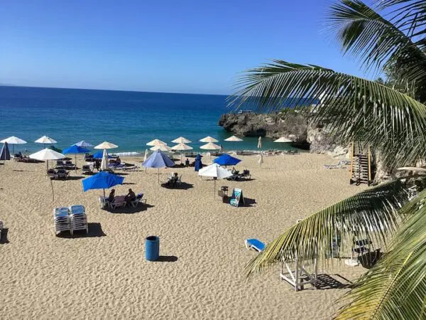 A tropical beach scene with multiple Ocean Front Lavish *Jr Penthouse* Exquisite Views, beach chairs, and people relaxing on the sand with a clear blue sky overhead and a rocky cove in the background.