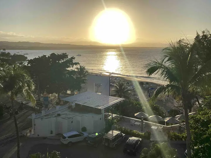 Sunset over a seaside town, with sun rays casting over the *Ocean Front Lavish Jr Penthouse* Exquisite Views, buildings, and palm trees, enhancing the reflective glimmer on the water.