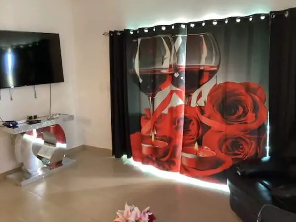 Ocean Front Lavish Jr Penthouse with large rose-printed curtain, flat-screen TV on wall, glossy white console, an LED-lit base, black sofa, and a small floral arrangement on the floor.