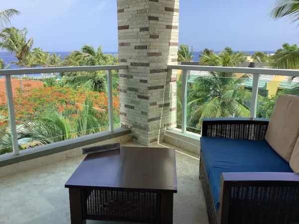 A balcony with two blue cushioned chairs and a coffee table, overlooking tropical trees and a glimpse of the Ocean Front Lavish *Jr Penthouse* Exquisite Views.