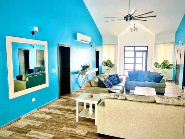 Bright living room with turquoise walls, modern furniture, ceiling fan, large windows, and a decorative mirror.