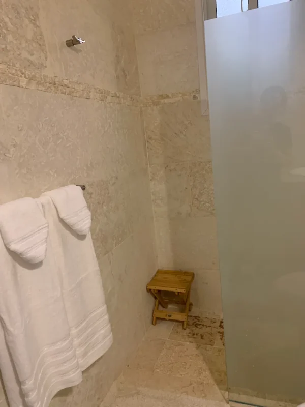 Interior of a bathroom featuring a glass shower door, hanging white towels, and a small wooden stool on tiled flooring.