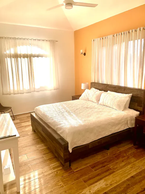 A bright, sunlit bedroom with a large wooden bed, white bedding, a ceiling fan above, sheer curtains on the windows, and a desk to the left.