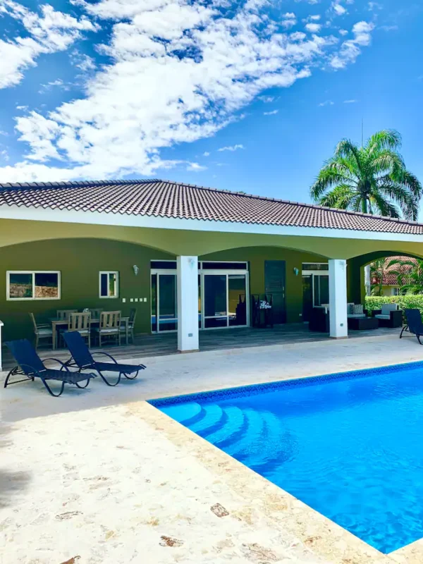 A tropical house with a swimming pool in the foreground, lounge chairs by the poolside, and a palm tree against a clear blue sky.