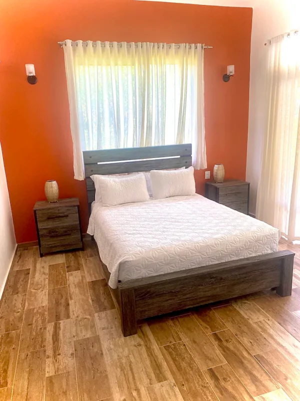 Bright bedroom with a queen-sized bed set against a vivid orange wall, white bedding, and two wooden nightstands, with sheer curtains on the windows.