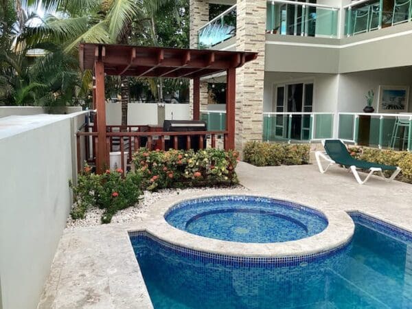 A tropical hotel courtyard featuring a small blue tiled Ocean Front Lavish Jr Penthouse hot tub, a wooden gazebo with bar seating, surrounded by lush plants and palm trees.