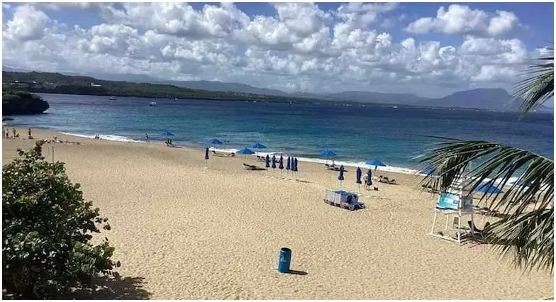 A scenic view of a sandy beach with blue umbrellas, clear sky, and calm sea in the background, flanked by lush greenery and beachfront villas.